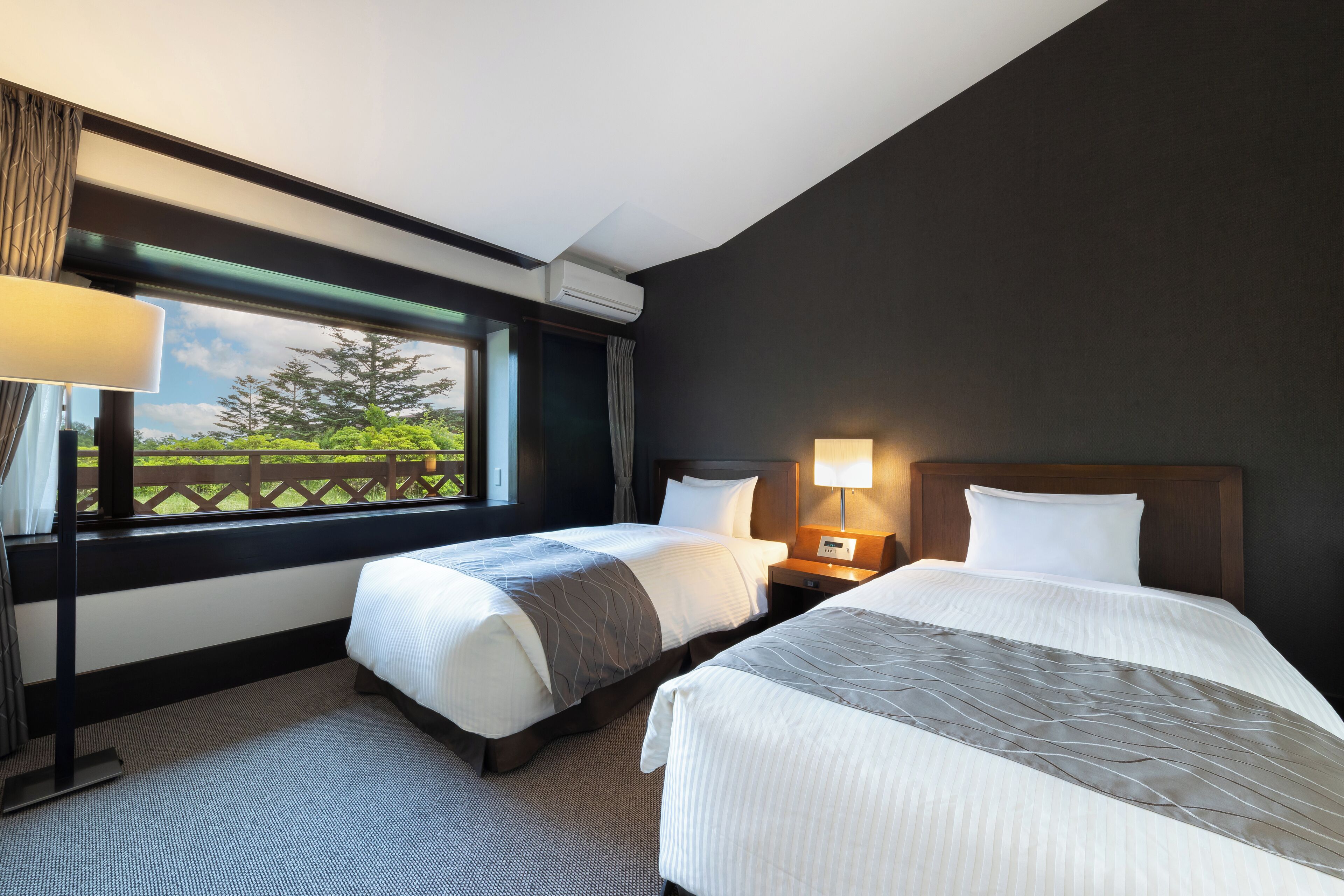 A hotel room with two beds, a window with a view of greenery, an air conditioning unit, and a nightstand with a lamp.