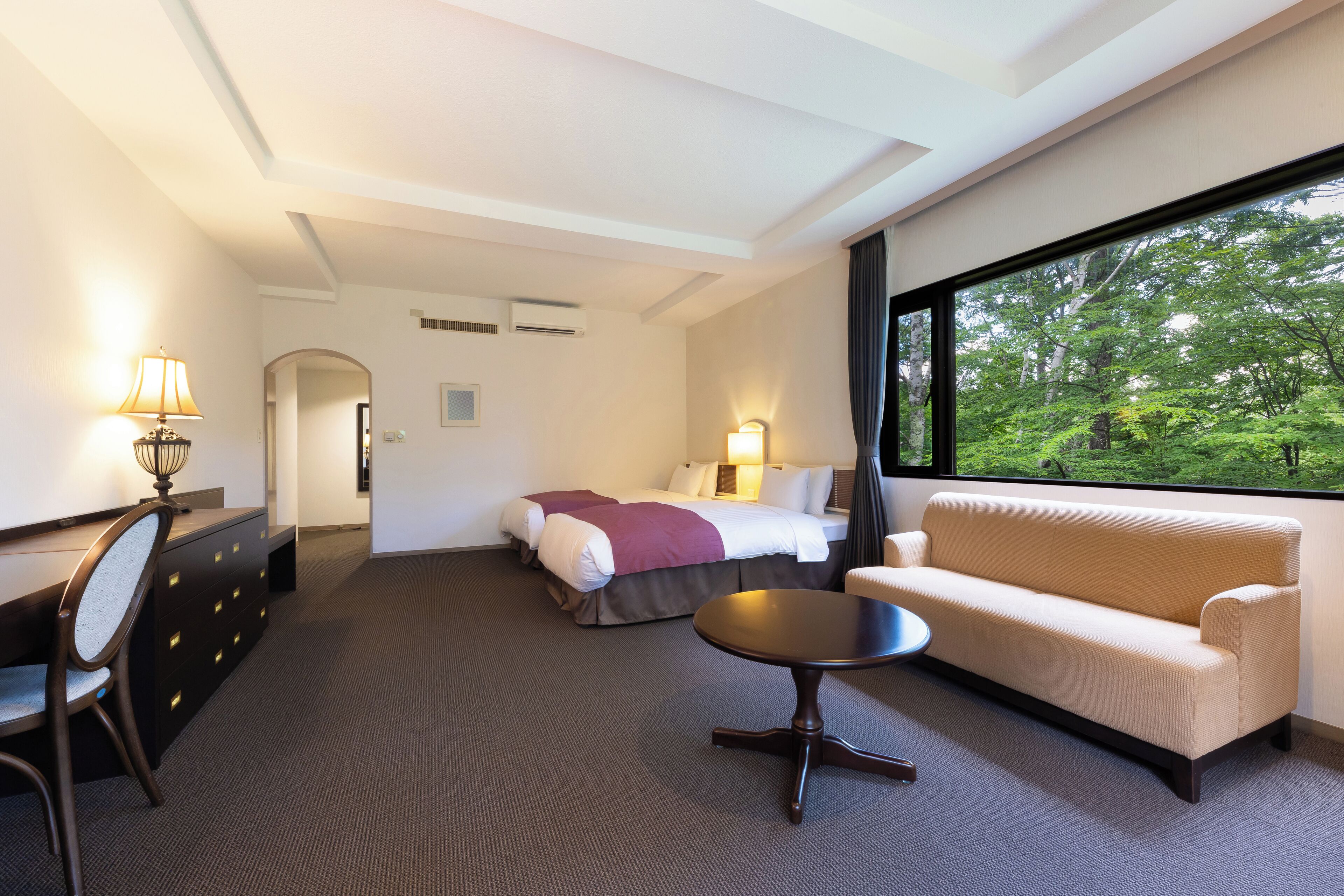 A hotel room with a bed, sofa, desk, and a view of greenery through a large window.