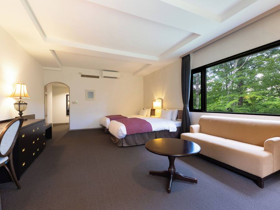 A hotel room with a bed, sofa, desk, and a view of greenery through a large window.