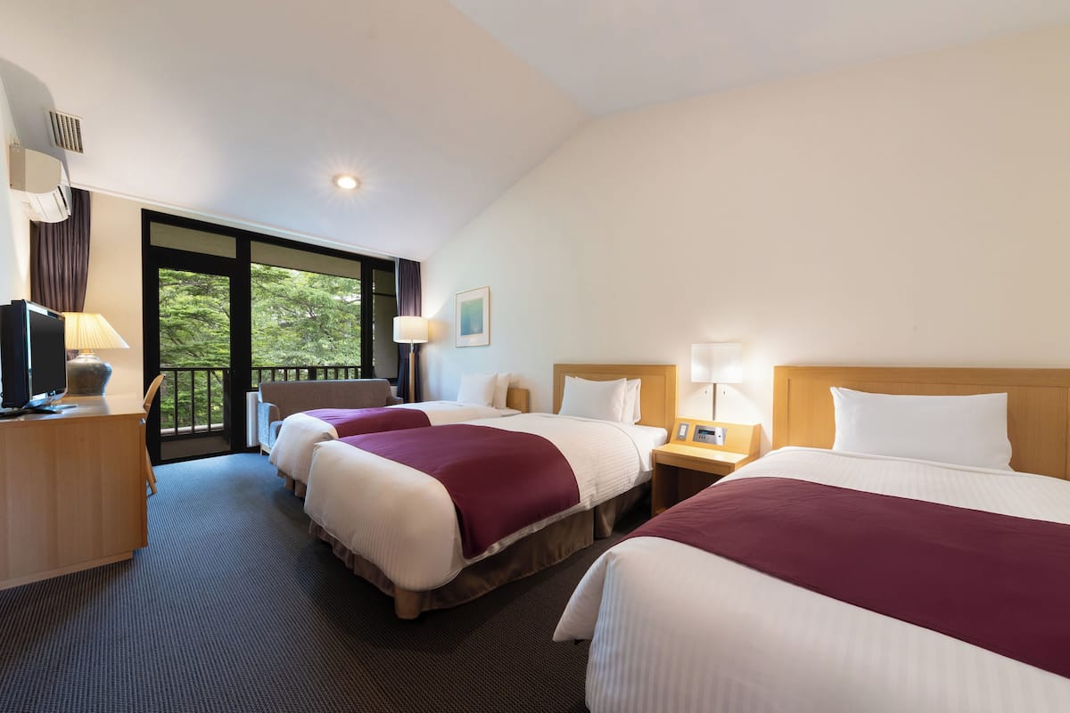 A hotel room with three beds, a desk with a computer, a TV, and a balcony with a view of trees.