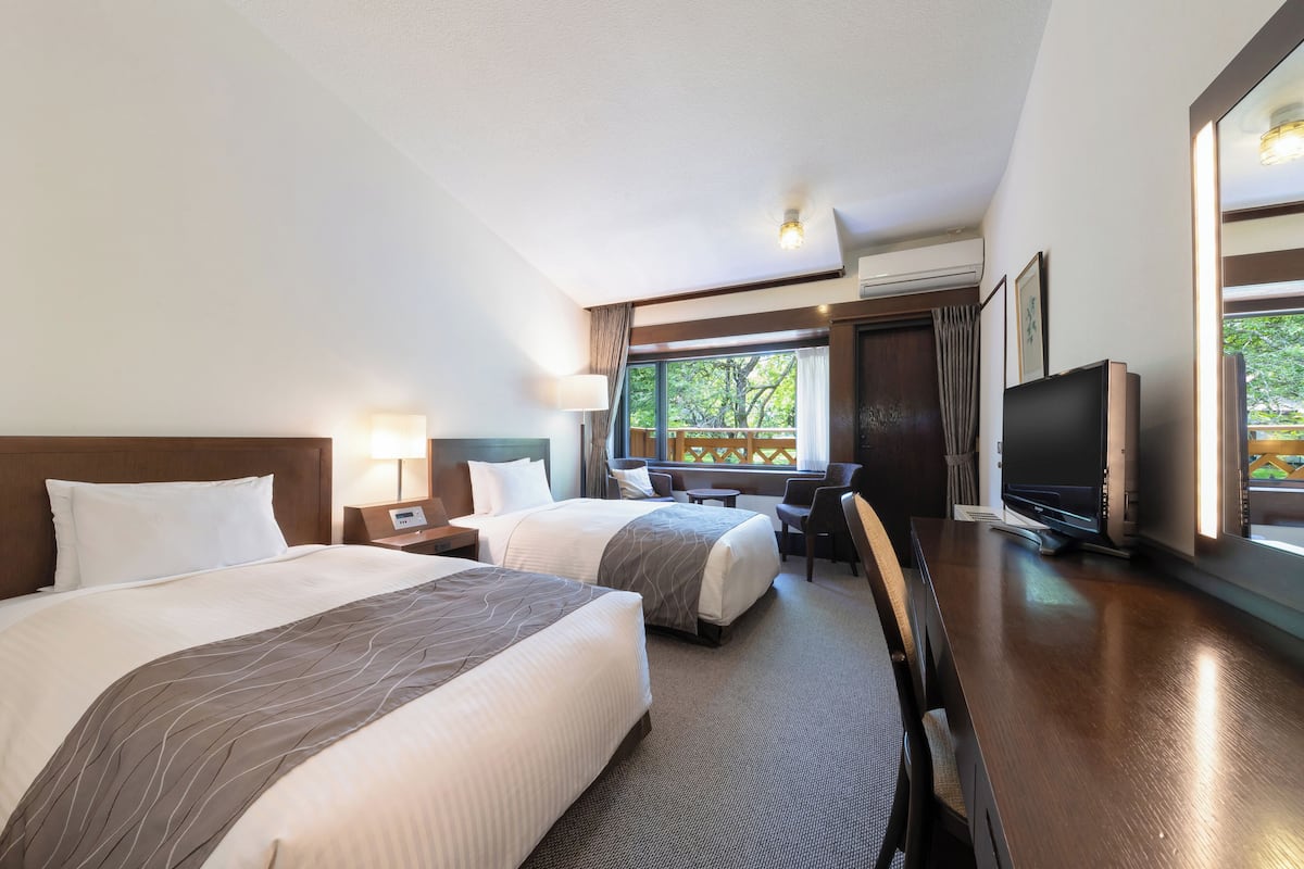 A hotel room with two beds, a desk, a chair, a TV, and a window with a view of trees.