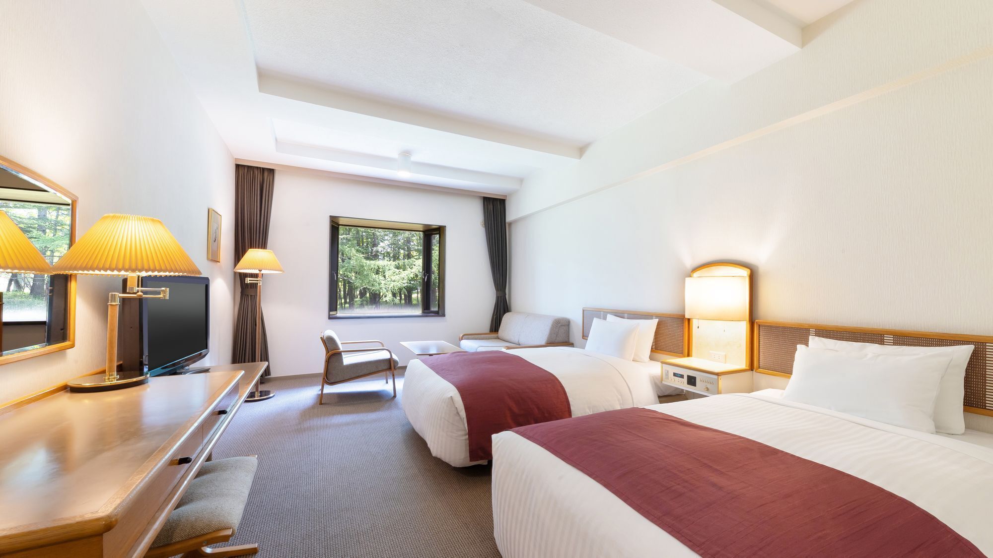 Ein Hotelzimmer mit zwei Betten, einem Schreibtisch, einem Sessel, einem Fernseher und einem Fenster mit Blick auf Bäume.