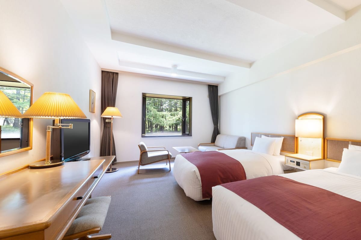 A hotel room with two beds, a desk, a chair, a TV, and a window with a view of trees.
