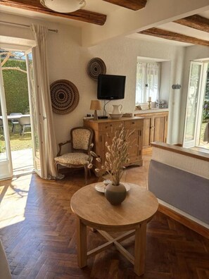 Una habitación con piso de madera, una mesa redonda con un florero, una silla, un televisor y una cocina con armarios y ventana.