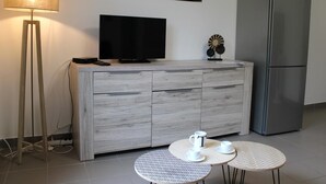En moderne stue med et gråt skab, et fladskærms-tv, en gulvlampe, et køleskab og to små runde borde med kopper.