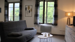 En stue med en grå sofa, et rundt sofabord med kop og underkop, en radiator og et fjernsyn på et træskab.