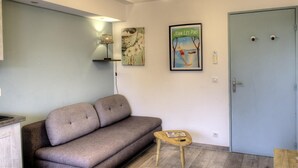 A room with a grey sofa, a small wooden table, a framed picture, a light blue door, and a wall-mounted lamp.