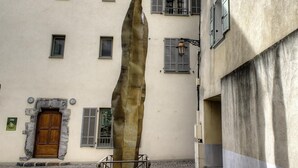Ein Gebäude mit einer großen, einzelnen Steinskulptur davor, einer Holztür mit Steinumrandung und mehreren Fenstern mit Fensterläden.