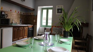 A dining area with a green table set for four, a potted plant, and a kitchen in the background. - La Maison Commune - CAILLE (CAILLE)