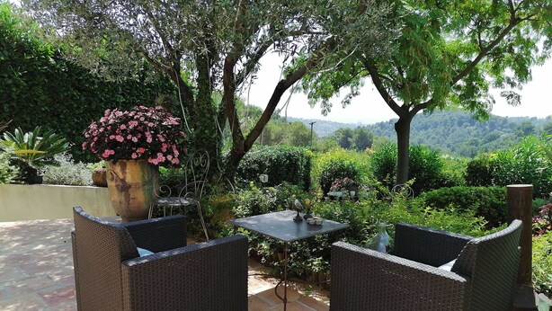 An outdoor patio with wicker furniture and a table, surrounded by lush greenery and a view of distant hills. - L'Olivier de St Paul (SAINT-PAUL-DE-VENCE)