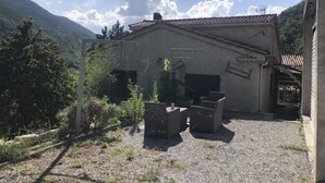 Un edificio rústico con techo de tejas, rodeado de grava y follaje, con montañas al fondo.