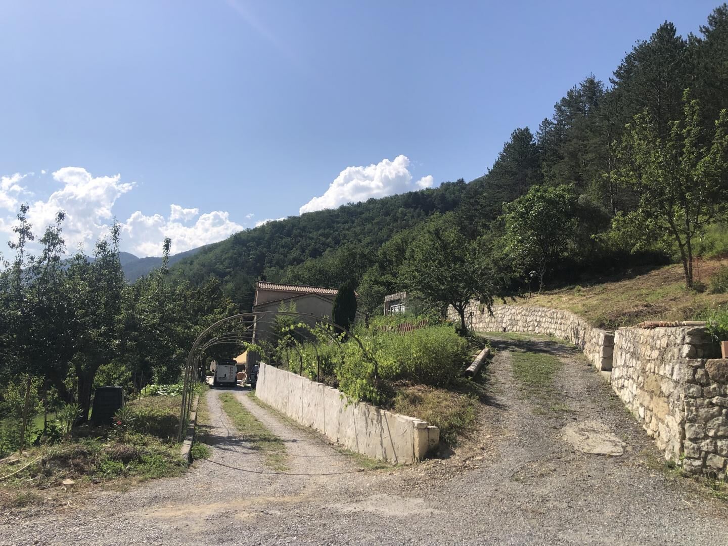 A rural road with a stone wall on one side and a building with a tiled roof in the distance.