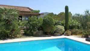 Una piscina con borde de baldosas, rodeada de plantas y árboles, y un edificio con techo de tejas al fondo.