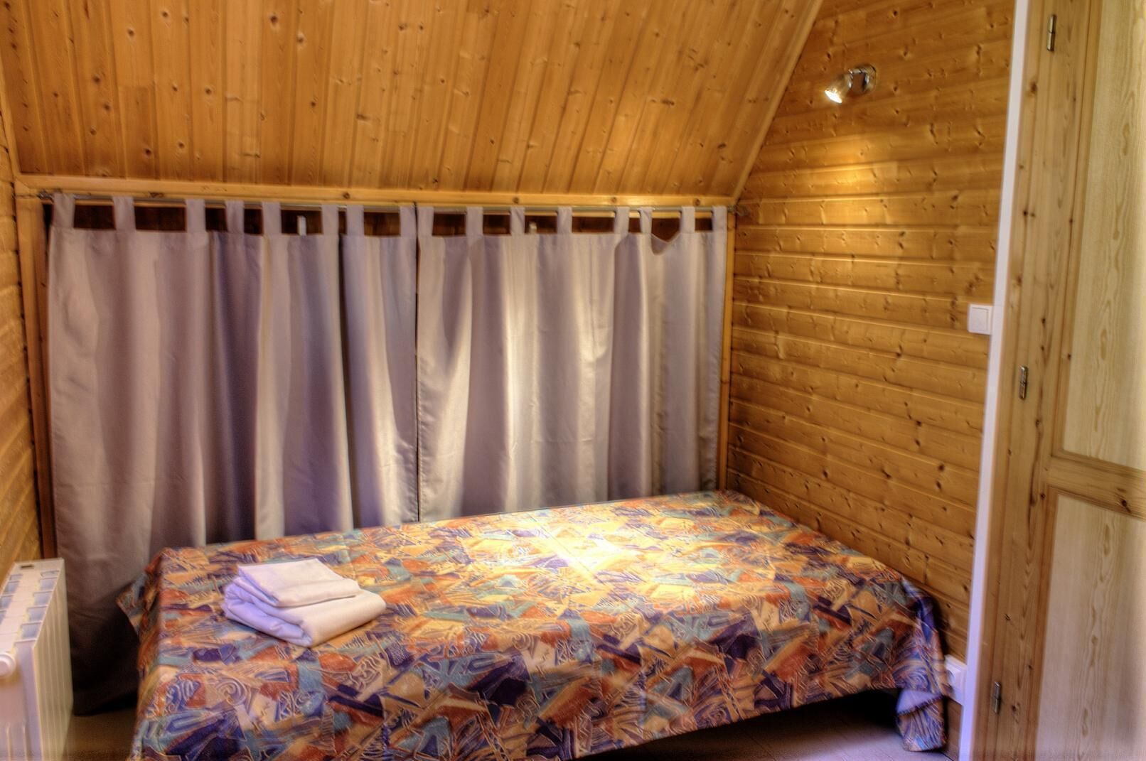 A small, cozy room with wooden walls and a sloped ceiling. It features a single bed with a colorful bedspread and a neatly folded towel.