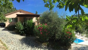 A house with a stone pathway leading to a pool area, surrounded by lush greenery and flowering plants.