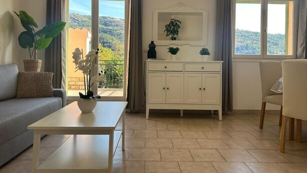 A living room with a sofa, a coffee table, a cabinet, and a chair. There are two windows with curtains, a view of greenery outside, and a potted plant on the windowsill.