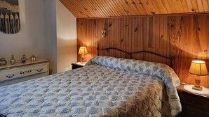 Un dormitorio con techo de madera, una cama con colcha estampada, una mesita de noche con lámpara y un aparador con adornos.