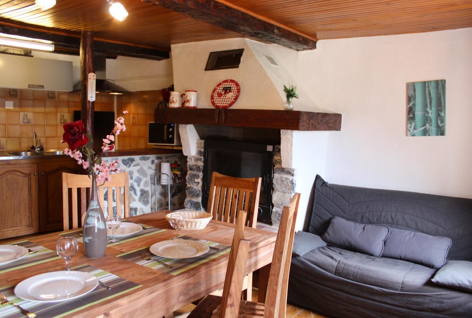 Un coin repas chaleureux avec une table en bois dressée pour quatre personnes, une cheminée et un canapé gris.