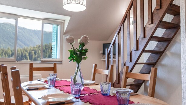 Une table à manger est dressée avec des assiettes, des verres et un vase rempli de fleurs, dans une pièce dotée d’un escalier en bois et offrant une vue sur les montagnes par la fenêtre.