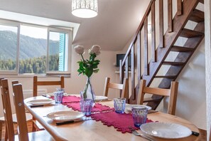 A dining table set with plates, glasses, and a vase with flowers, in a room with a wooden staircase and a view of mountains through the window. - Le Génépi - ROUBION - Gîte (ROUBION)