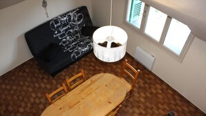Un coin repas avec une table et des chaises en bois, un canapé noir recouvert d’un tissu à motifs, un luminaire suspendu et une fenêtre avec des stores.