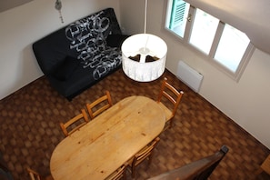 A dining area with a wooden table and chairs, a black sofa with a patterned cover, a hanging light fixture, and a window with blinds. - Le Génépi - ROUBION - Gîte (ROUBION)