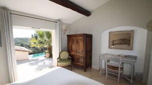 A bedroom with a bed, a desk, a chair, and a wardrobe. There is a balcony with a view of trees and a pool. - Cueille la Nuit - TOURRETTES-SUR-LOUP (TOURRETTES-SUR-LOUP)