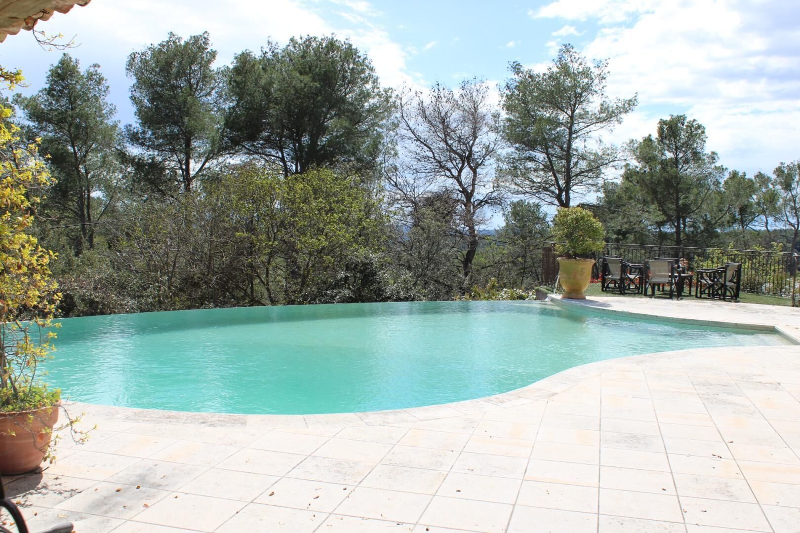 A swimming pool with a tiled deck, surrounded by trees and a patio area with outdoor furniture.