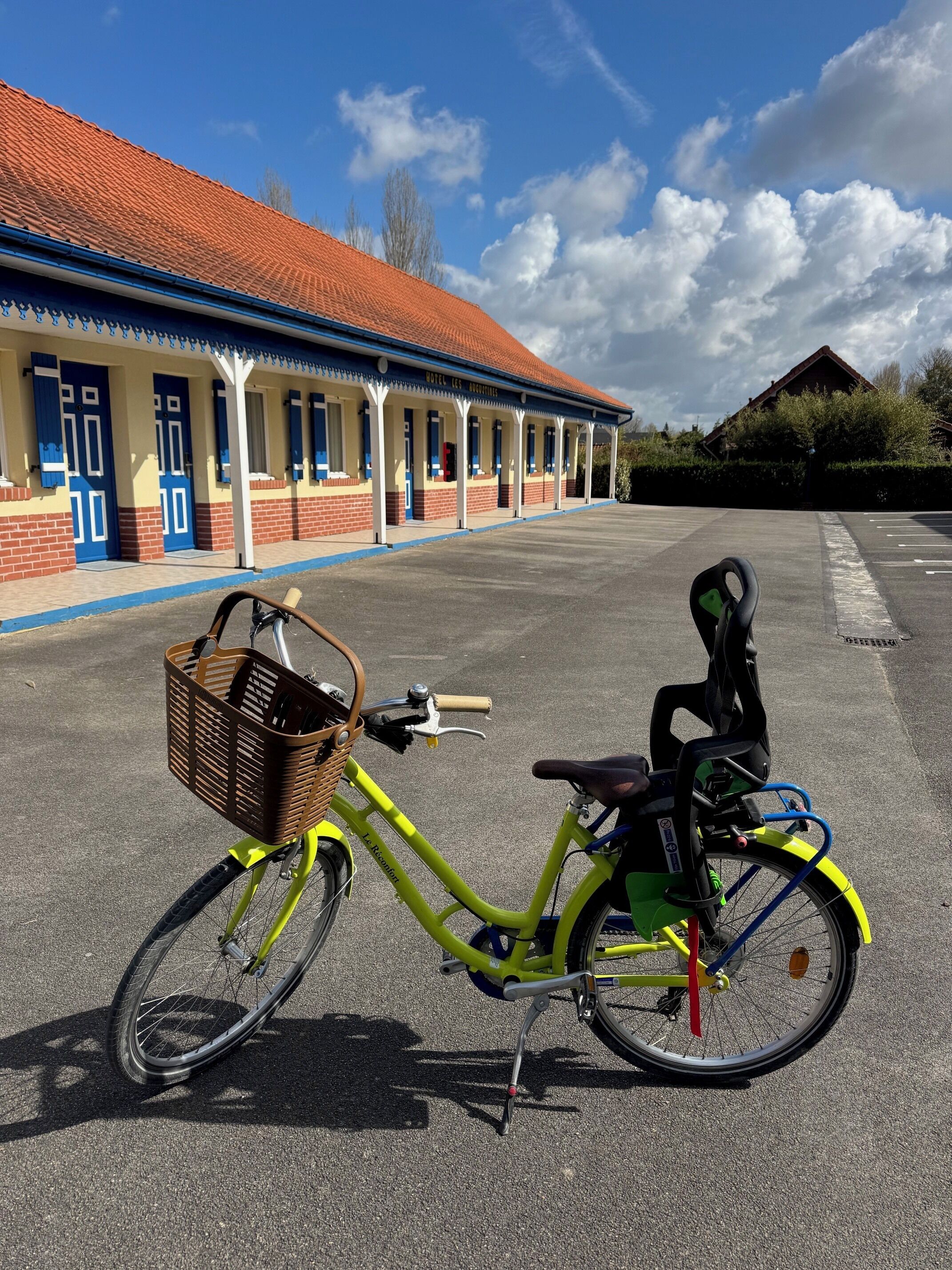 Un vélo jaune, muni d’un panier et d’un fauteuil roulant fixé à l’arrière, est garé devant un immeuble avec un toit rouge et des portes bleues.