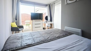 A bedroom with a bed, a desk, a chair, a television, and a window with curtains. - Hotel Les Augustines (Quend)