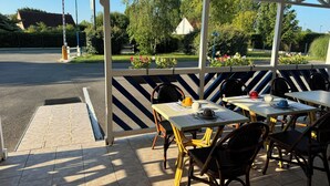 An outdoor seating area with tables and chairs, a covered walkway, and a sign indicating a hotel. - Hotel Les Augustines (Quend)