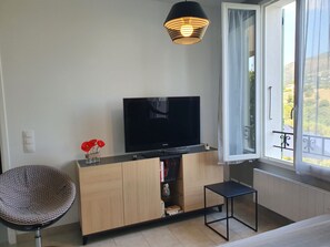 A room with a television on a wooden cabinet, a chair, a small table, and a window with a view of greenery. - Le Magnolia - Colomars (Colomars)