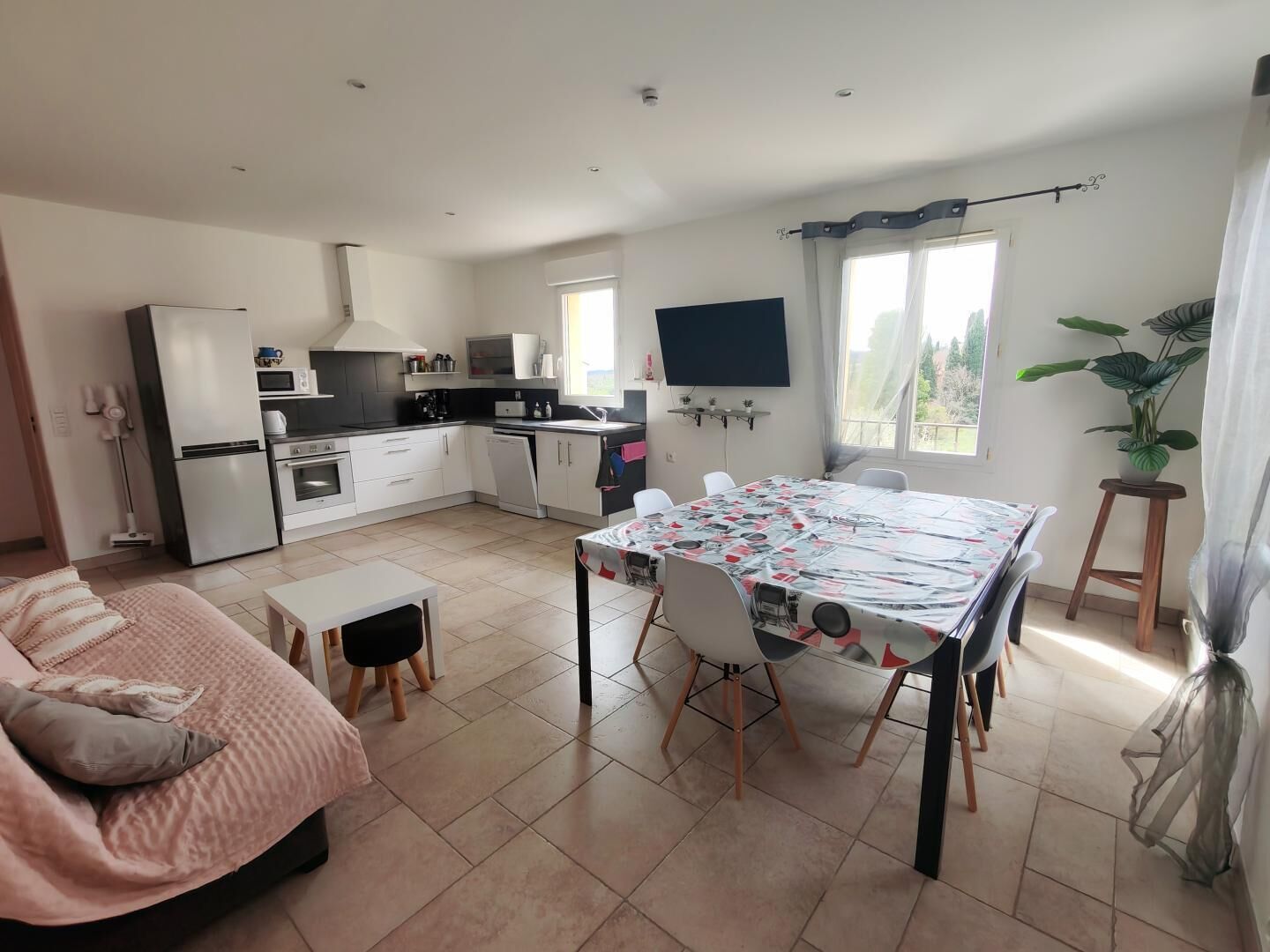 Una cocina moderna con armarios blancos, una mesa de comedor con sillas, un televisor montado en la pared y un amplio ventanal con cortinas.