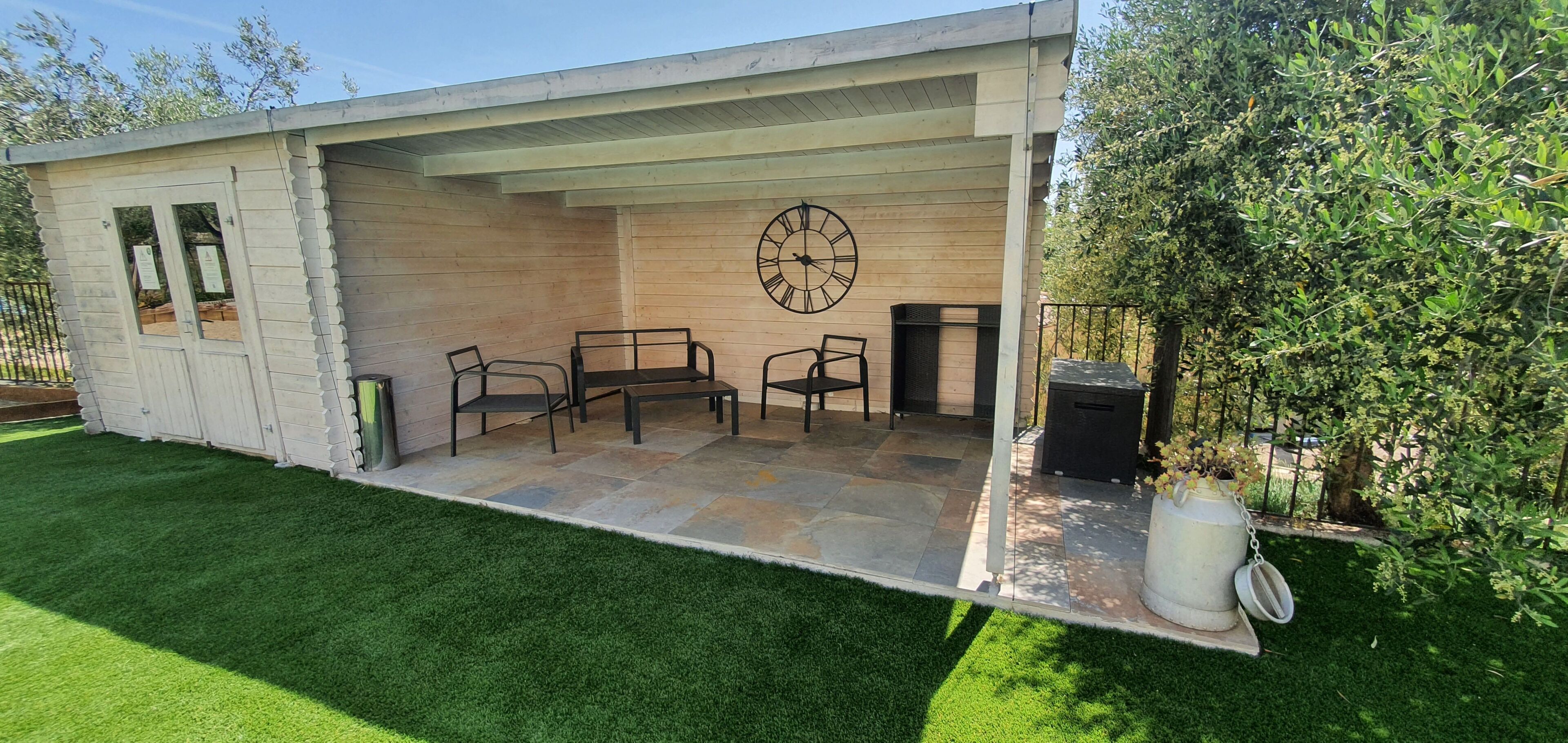 A covered patio with a table and chairs, a clock on the wall, and a trash can.
