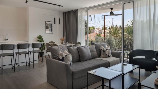 Een moderne woonkamer met een grijze bank, een eethoek met stoelen, een kleine tafel en uitzicht op palmbomen door grote ramen.