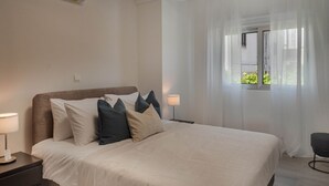 Een slaapkamer met een groot bed, een nachtkastje, een lamp en een raam met dunne gordijnen.
