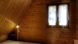 A small, cozy room with wooden walls and a sloped ceiling. There is a single bed with a white sheet and two folded towels. A window with white curtains allows natural light to enter.