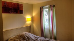 A bedroom with a bed, a wall-mounted lamp, and a window with striped curtains. - Le Balcon - PEILLE - Gîte (PEILLE)