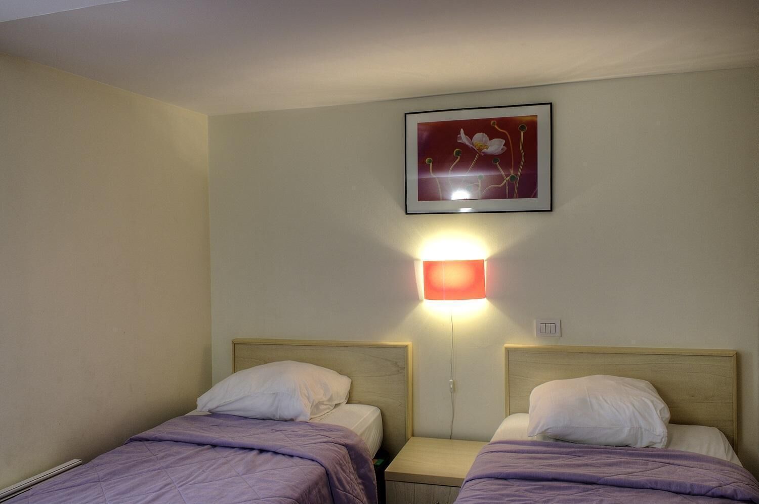 A hotel room with two single beds, a nightstand with a lamp, and a framed picture on the wall.