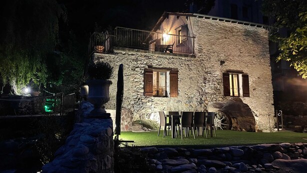 A stone building with a balcony, a dining area with chairs, and a stone wall in the foreground.