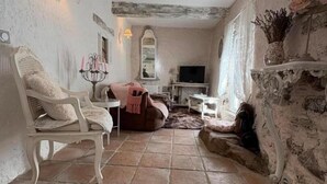 A rustic living room with exposed beams, a stone floor, a white armchair, a television, and a fireplace.