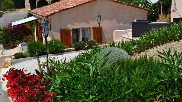 A small, single-story building with a tiled roof, surrounded by a well-maintained garden with various plants and flowers. - Little House of Friends (LA GAUDE)