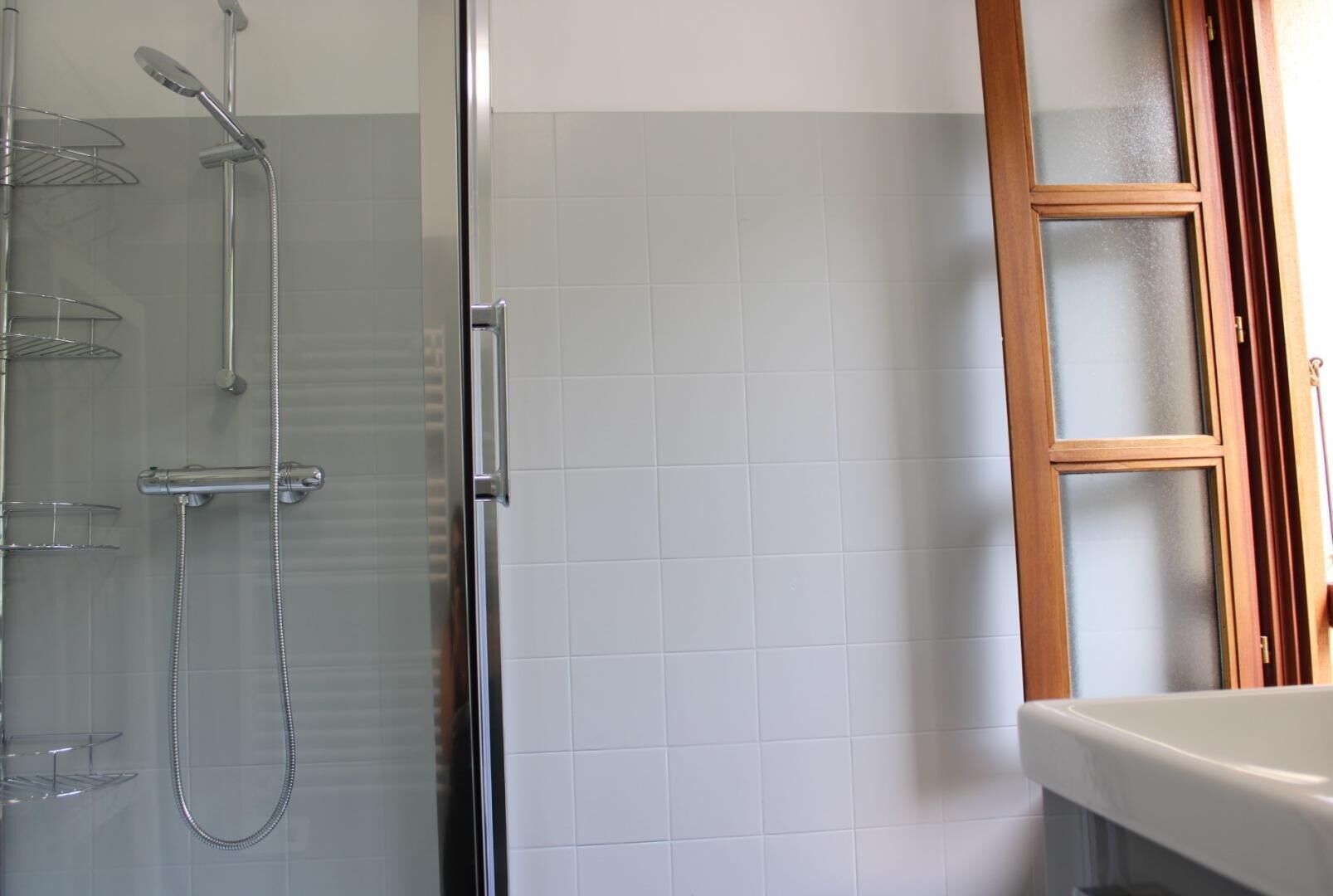 A bathroom with a glass shower enclosure, a wooden-framed window, and a white sink.