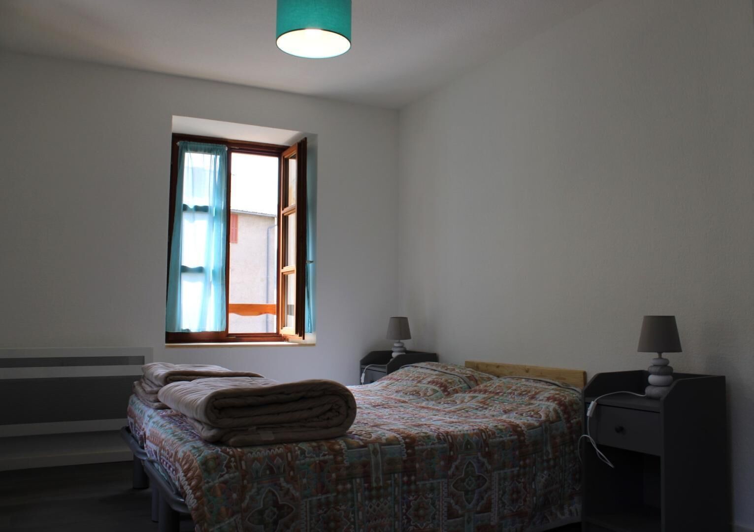 A bedroom with a bed, a window with curtains, a bedside table with a lamp, and a desk with a lamp.