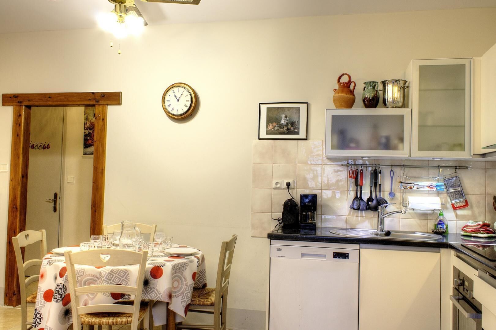 A compact kitchen with a dining area, featuring a clock, a coffee maker, and various kitchen utensils.