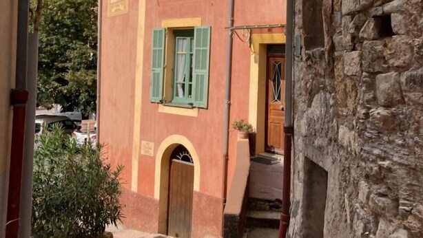 Un vicolo stretto con un edificio rosso, persiane verdi e un muro in pietra.