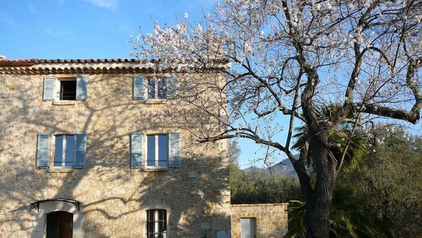 A stone building with multiple windows and a door, surrounded by greenery and a tree with blossoms. - Bastide de la Rousoulina (LE ROURET)
