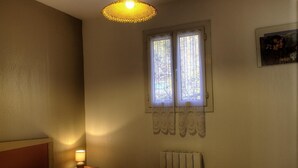 Un dormitorio con cama, una mesita de noche con lámpara, un radiador y una ventana con cortinas.