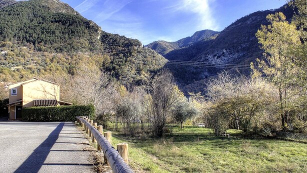 Un camino rural que lleva a un edificio con techo inclinado, rodeado de árboles y montañas.