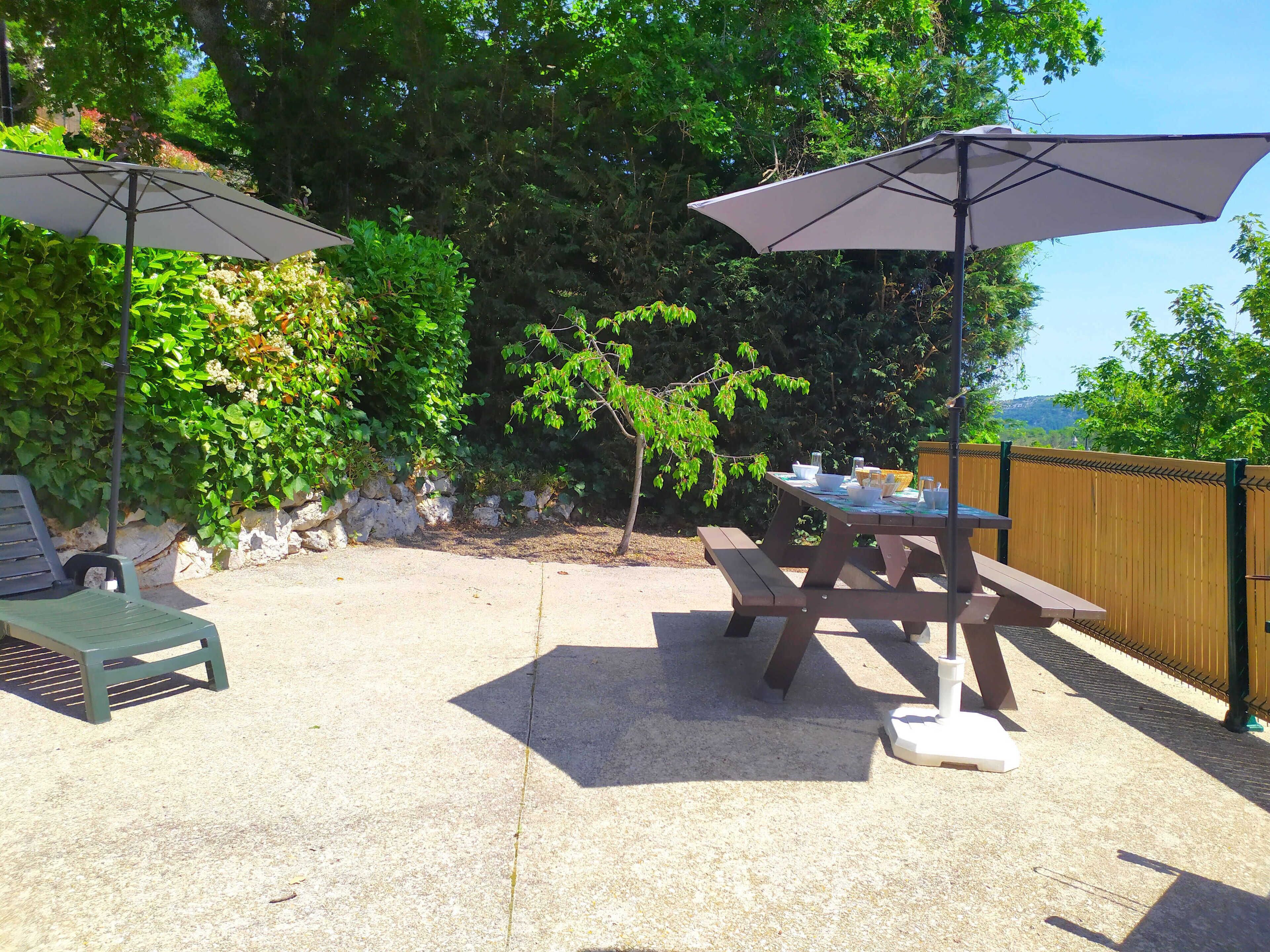 Une terrasse extérieure avec des tables de pique-nique et des parasols, entourée de verdure et d’une clôture en bois.